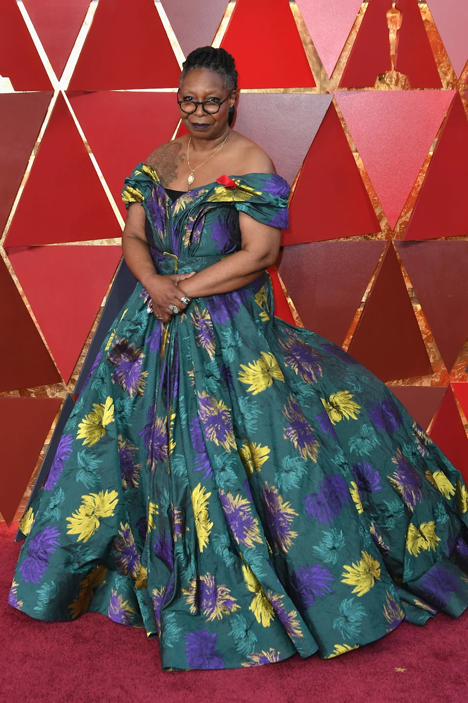 Person in an elegant off-shoulder gown with floral patterns stands on a red carpet, posing for the camera