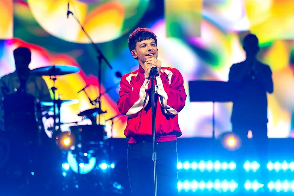 Singer on stage in a vibrant jacket performs with a colorful backdrop and band silhouettes