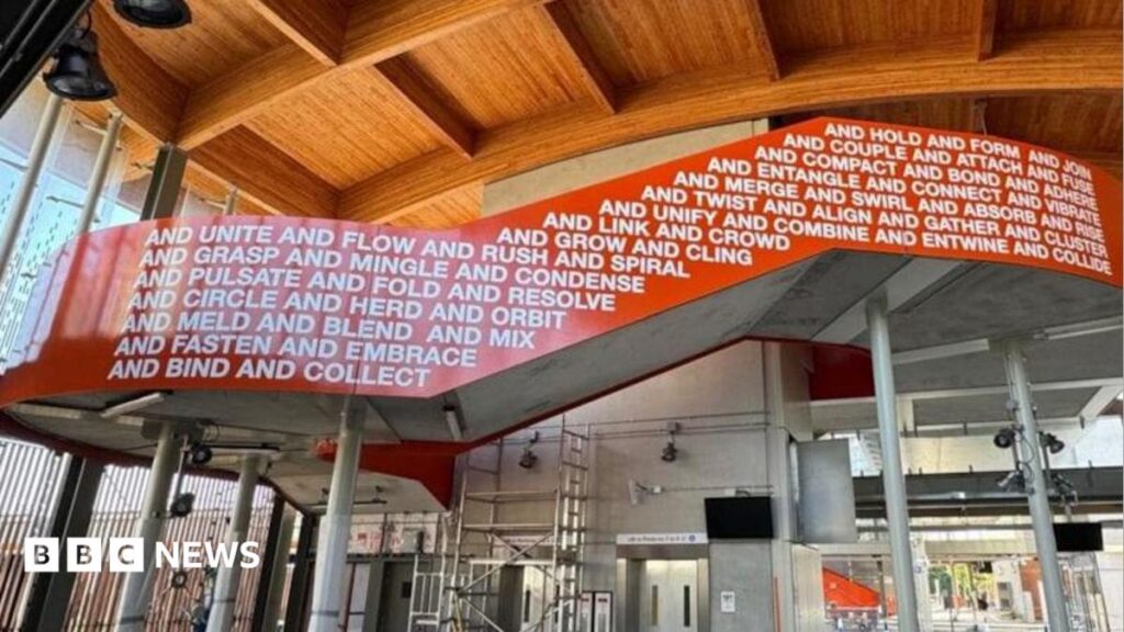 Cambridge South railway station unveils Turner prize nominee’s art The interior of a new railway station, which still has some scaffolding up as it is not yet complete. The ceiling is wooden and beneath is a stairway balustrade, sweeping from left to right, which is coloured bright orange and has words written in white on it.
