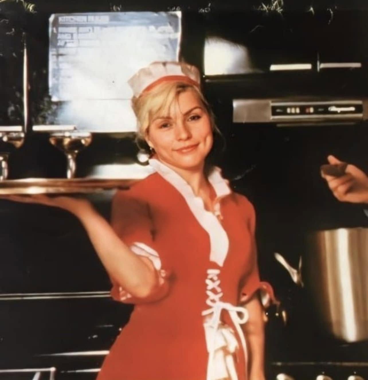A smiling woman in a red and white waitress uniform holds a tray with two glasses in a restaurant kitchen, standing in front of dark appliances and a large pot.