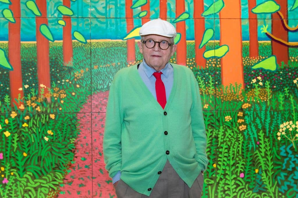 David Hockney stands in front of a colourful mural, wearing a green sweater and white cap.