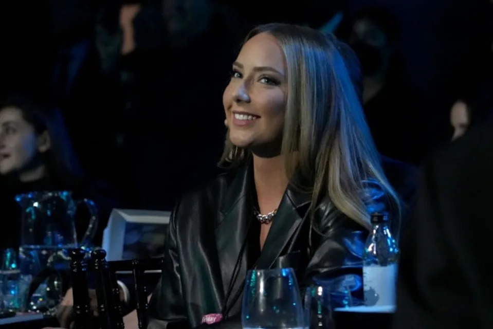 Woman sitting at an event in a chic leather jacket, smiling; guests are visible in the dimly lit background