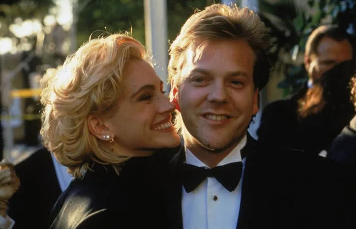 Two people in formal attire pose closely on a red carpet. One leans in, smiling widely; the other is in a tuxedo, smiling at the camera
