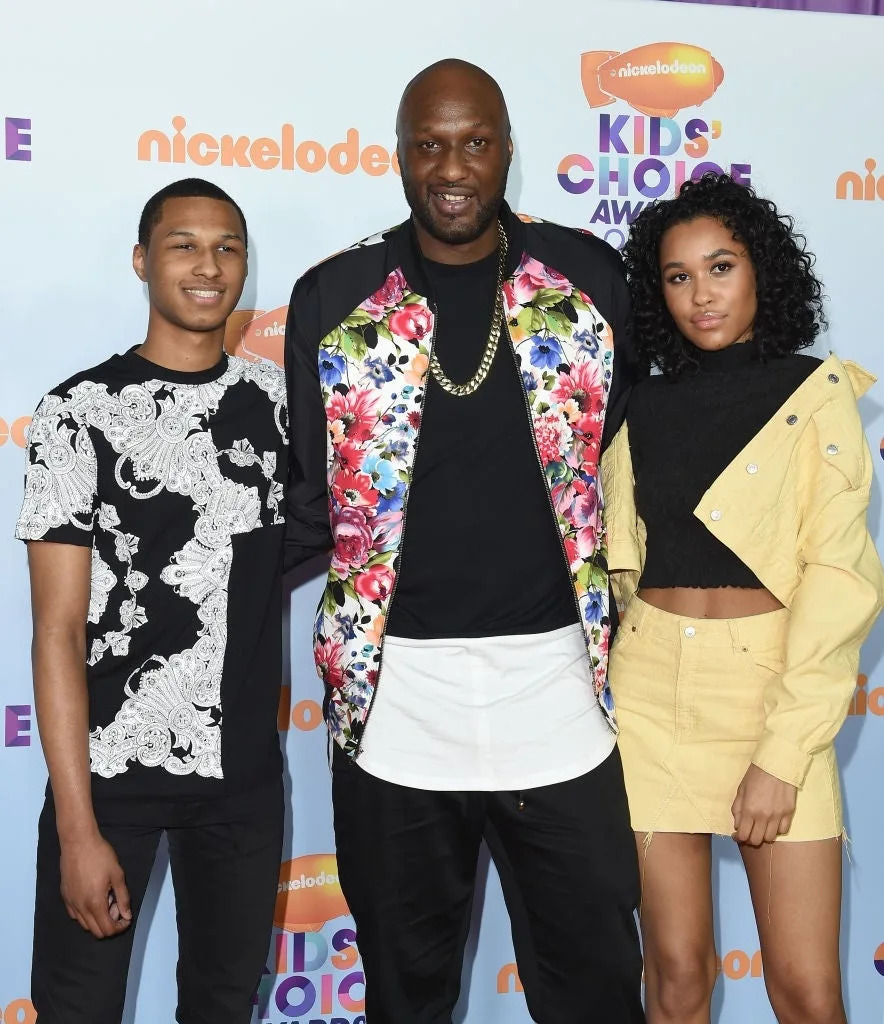 Three individuals pose at an event; the center person wears a floral jacket, while the others sport casual black-and-white and light-colored outfits