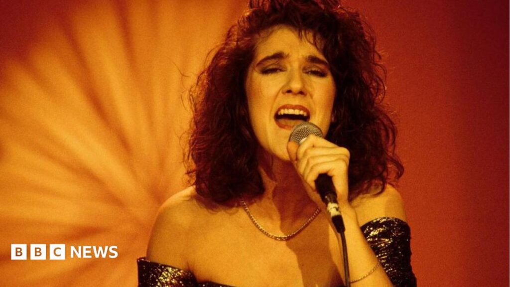 A young Celine Dion sings while holding a microphone. She is wearing a glittery strapless dress.