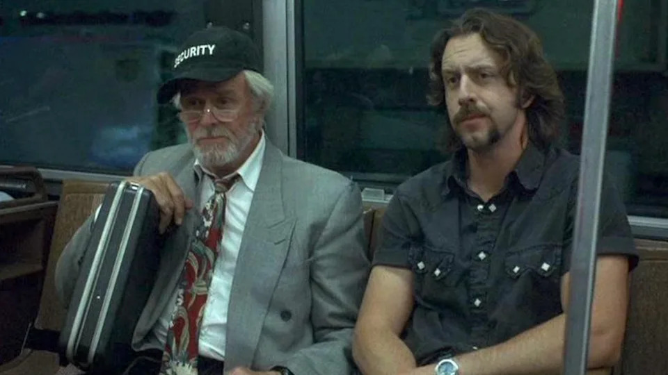 Two men sitting on public transport; one in a suit with a security cap and briefcase, the other in a casual shirt with a handlebar mustache