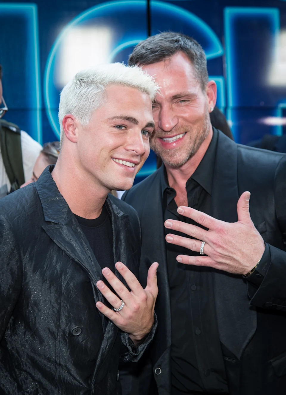 Two people smiling and showing rings, both wearing stylish dark suits, at an event.