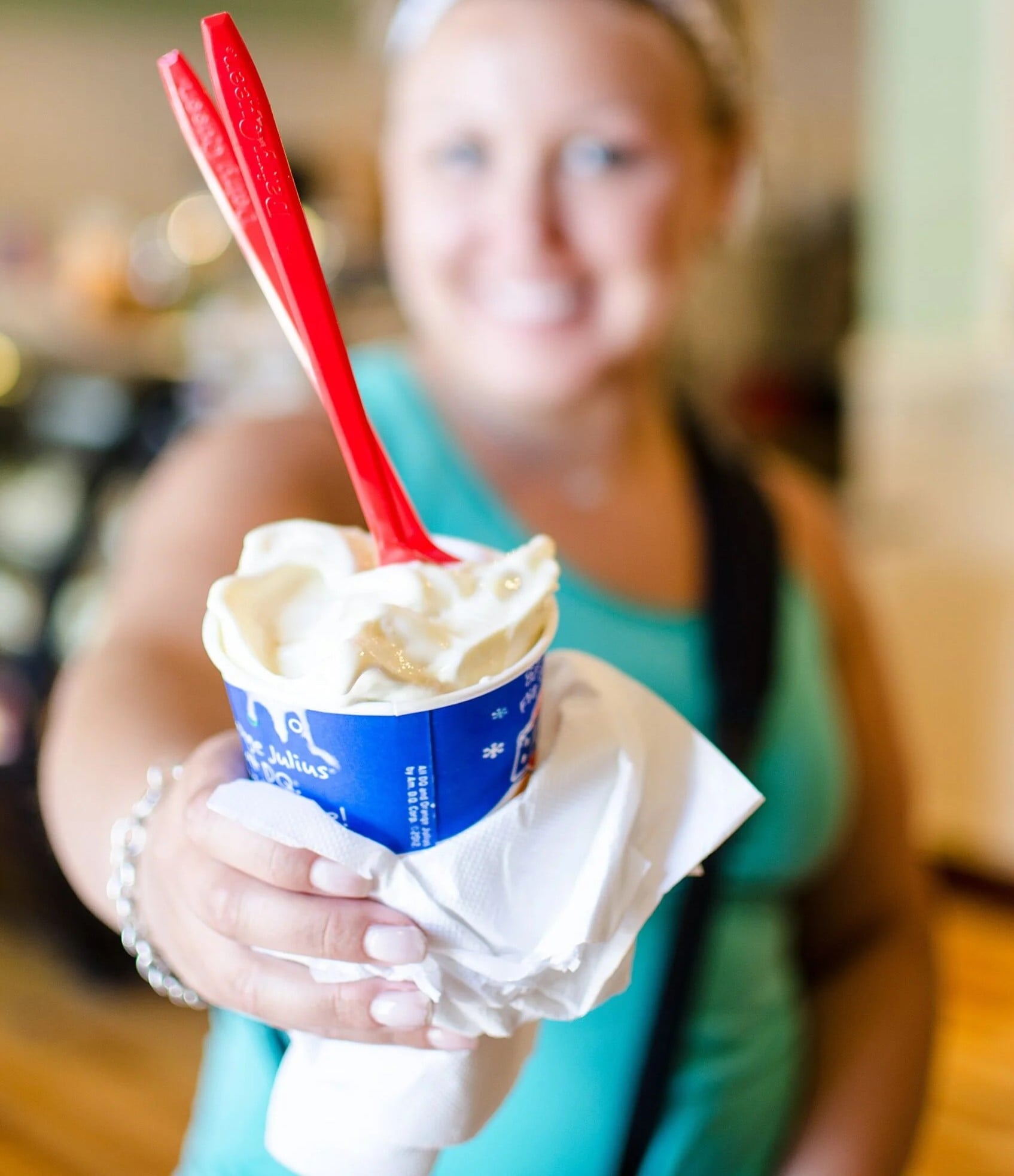 A person in a teal tank top smiles and holds out a cup of soft-serve ice cream with two red spoons. The cup is wrapped in a napkin, and the background is blurred.