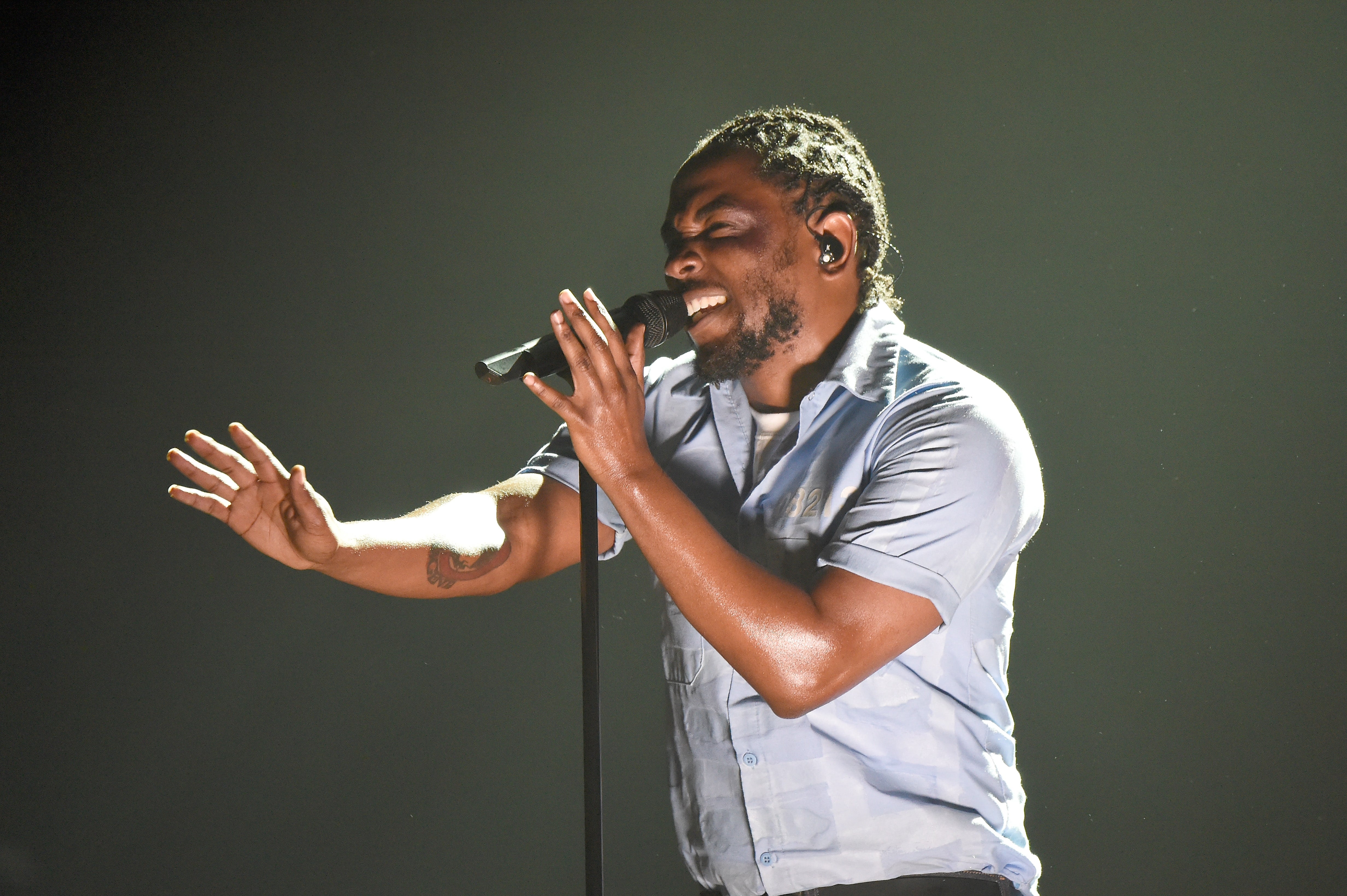 Kendrick Lamar passionately singing into a microphone on stage, wearing a short-sleeve shirt