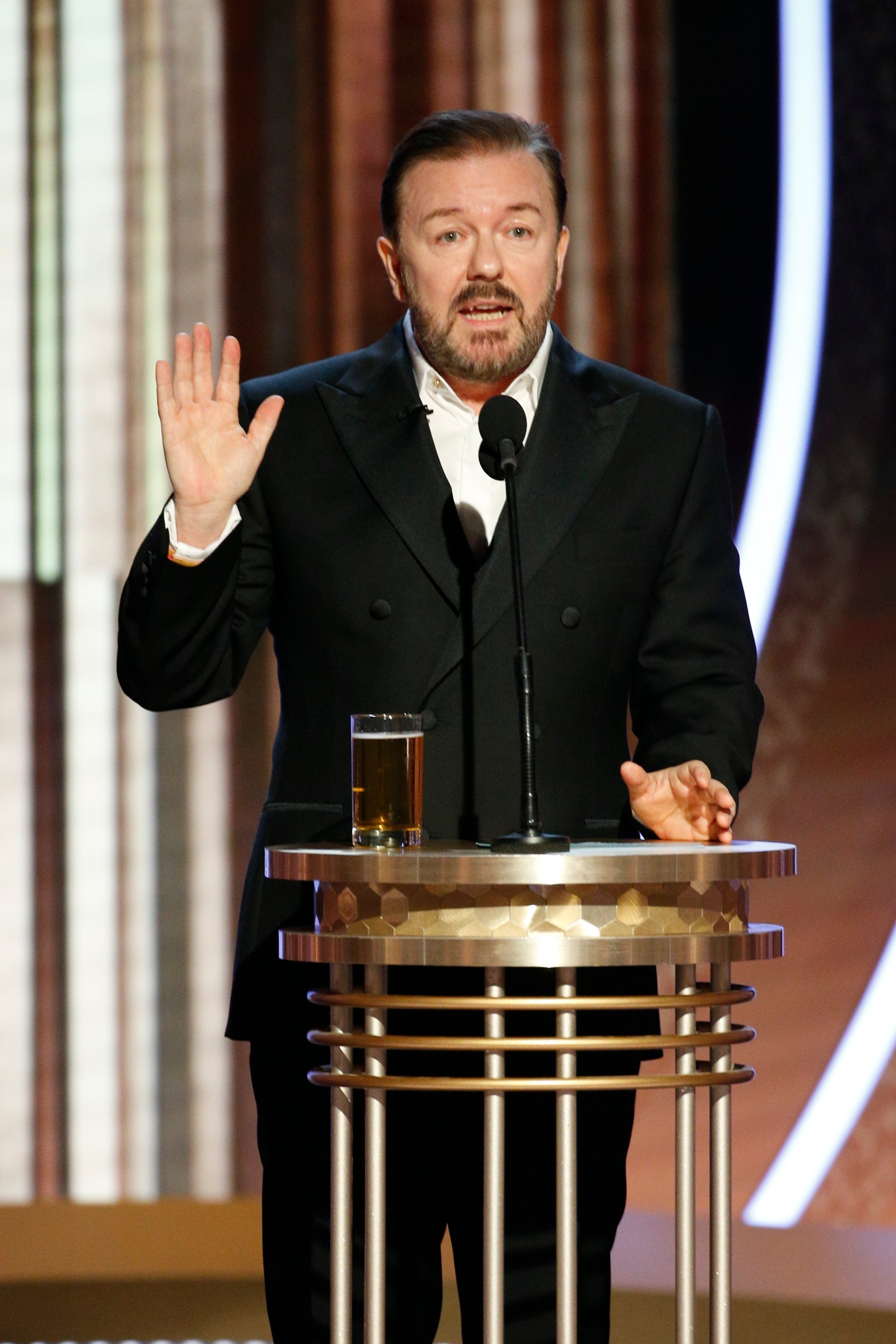 Ricky Gervais in a tuxedo speaks at a podium with a hand raised, holding a drink