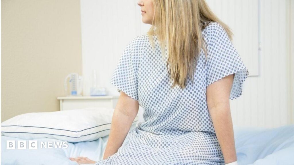 Taboos are on the table at Bury St Edmunds women’s health event A woman wearing a white and blue hospital gown sits on the edge of a patient bed with her legs hanging down and her hands resting on the edge. She is looking to the left. The image is cut off above her nose. She has long, straight blonde hair.