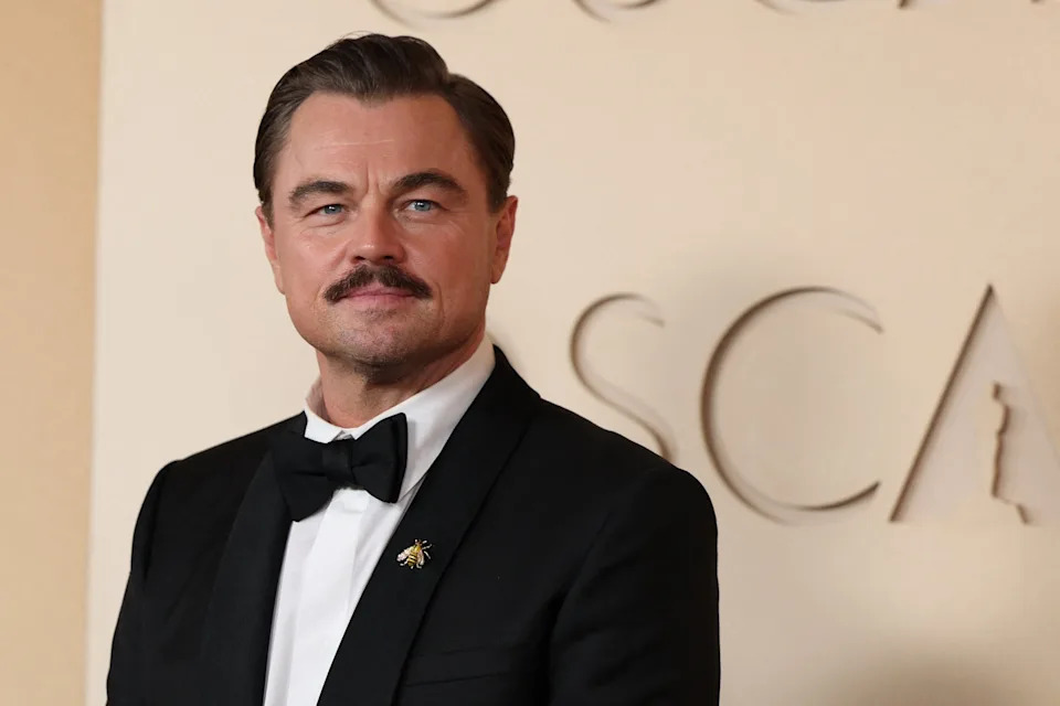 A person in a tuxedo with a bow tie and a lapel pin, standing against a neutral backdrop