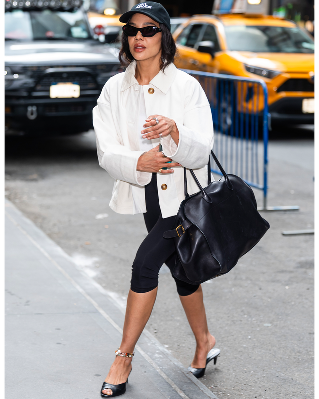 Person walking in city wearing white coat, black hat, sunglasses, and carrying a large black bag. Person walking in city wearing white coat, black hat, sunglasses, and carrying a large black bag.
