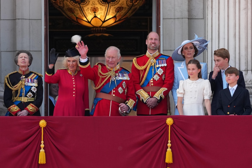 The British royal family in official dress stand on a balcony waving to a crowd.