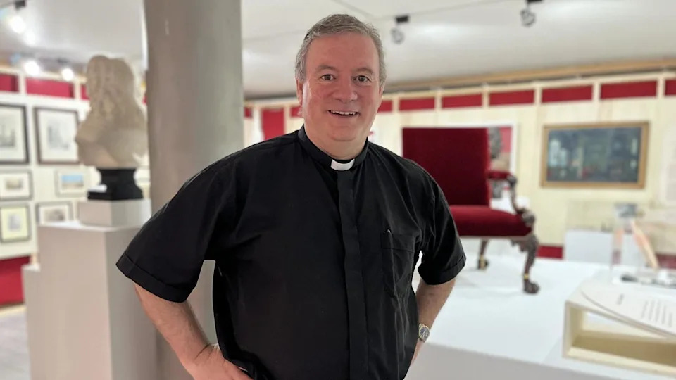 The Reverend Sam Wells wearing a black shirt and do collar standing within the exhibition with a chair, a bust of a head and paintings in the background