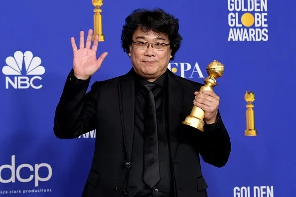Bong Joon-Ho in a black suit holding a Golden Globe award, smiling, and waving at a Golden Globe Awards event