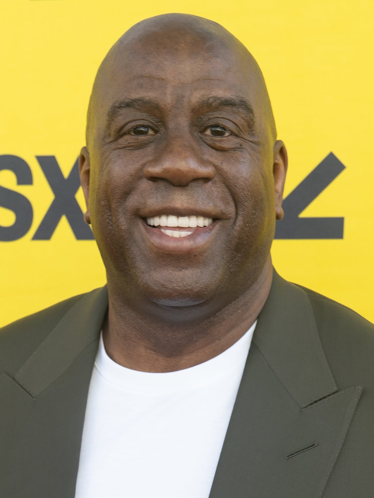 A man with a clean-shaven head, broad smile, and dark blazer over a white shirt stands in front of a yellow background with black text.
