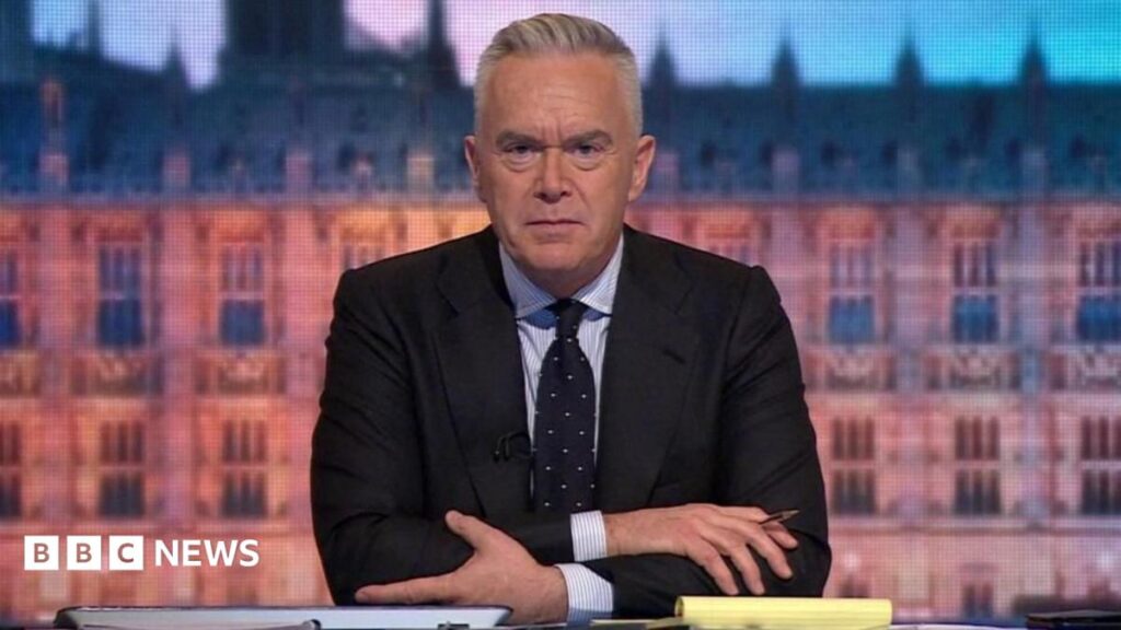A TV anchor white hair wearing a dark suit and tie. Behing him is an image of a brown building. He has his arms folded on a desk, is holding a pen and is looking directly into the camera.