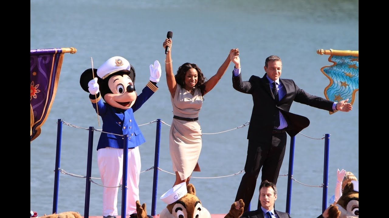 Three people stand on a stage by the water, including a person in a Mickey Mouse costume dressed as a captain, a woman holding a microphone, and a man in a suit. They hold hands and raise arms in celebration.