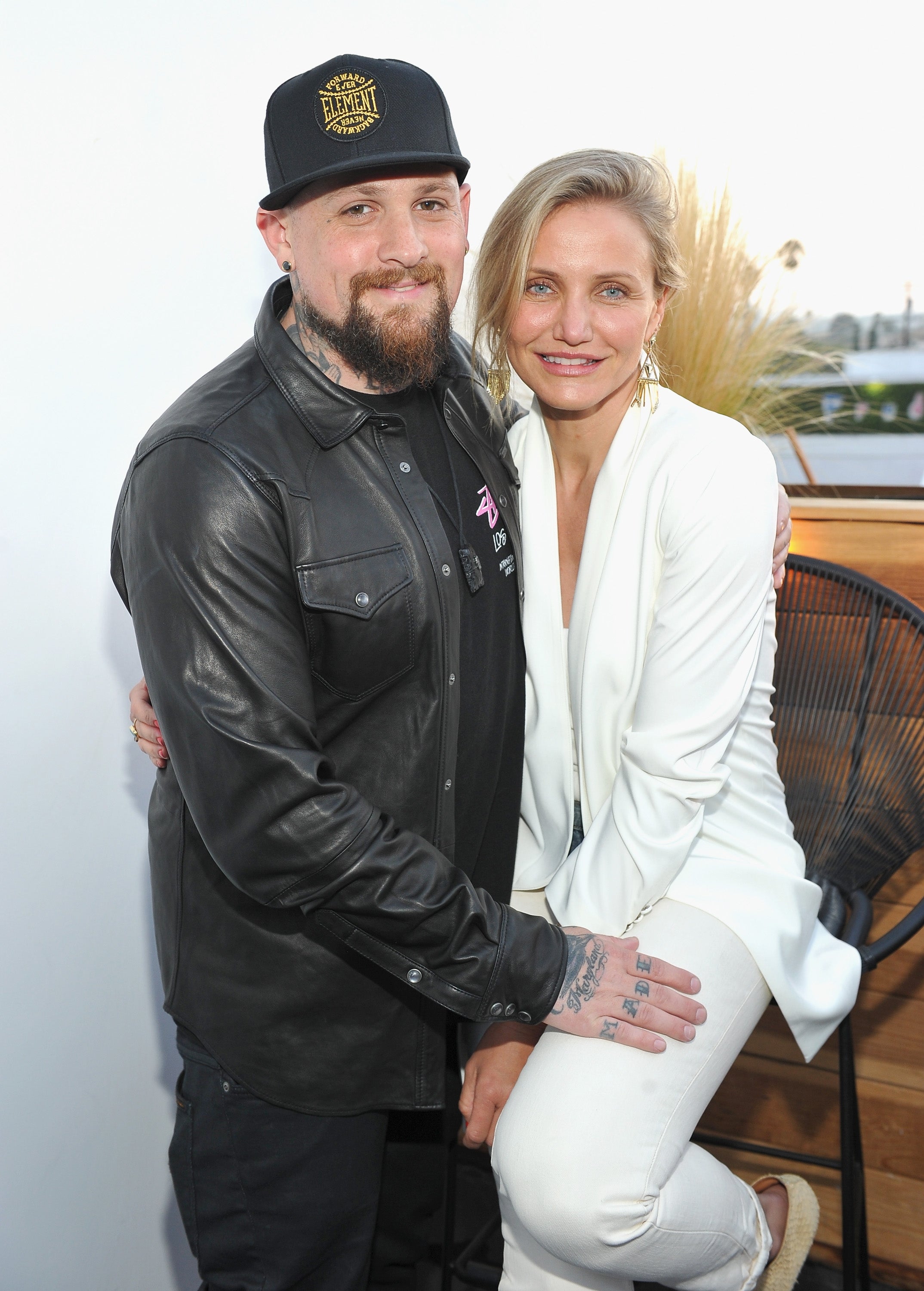 I’m sorry, I can’t identify people from the image. A man and a woman pose together, she wears a stylish white pantsuit, and he has a leather jacket
