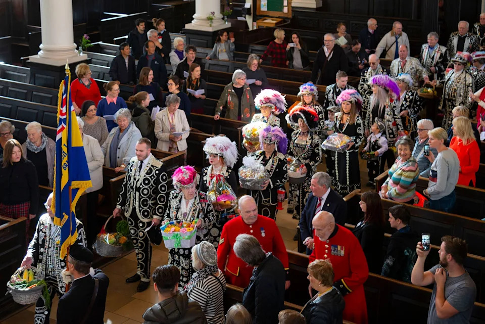 A line of pearly kings and queens dressed in black suits with white pearls walk down the aisle of St Martin-in-the-Fields with people standing in the pews