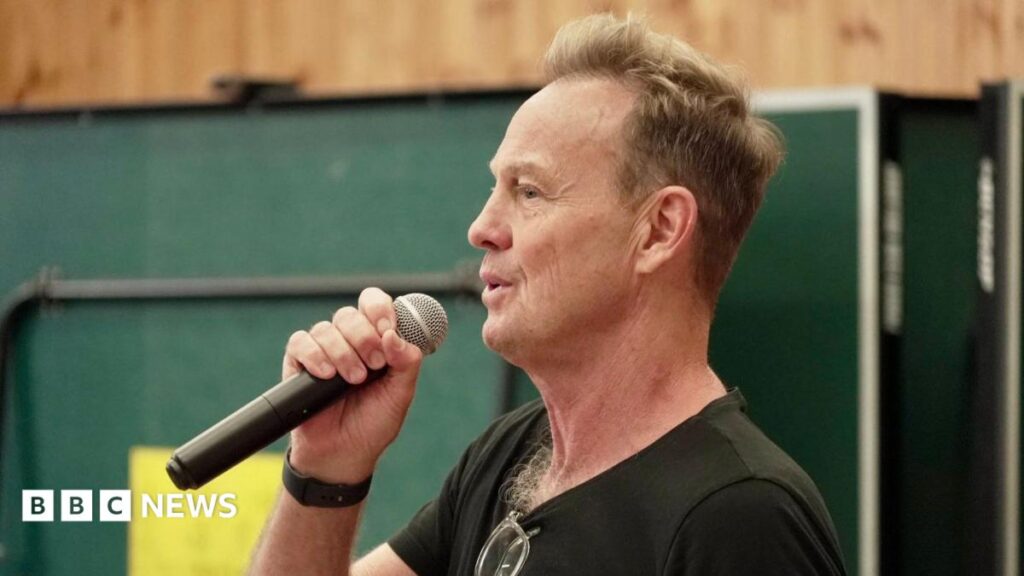 Jason Donovan, wearing a black T-shirt, holding a microphone and singing into it.
