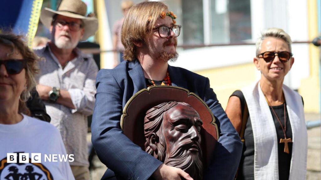 ‘Severed head’ paraded at start of Penzance’s Golowan Festival A man with a ring of orange flowers in his hair carries a wood head of St John during a parade. He is wearing a blue suit jacket. He also has glasses on and has straight brown hair and a brown goatee beard. Two women are stood either side of him and a man wearing a hat is behind him.
