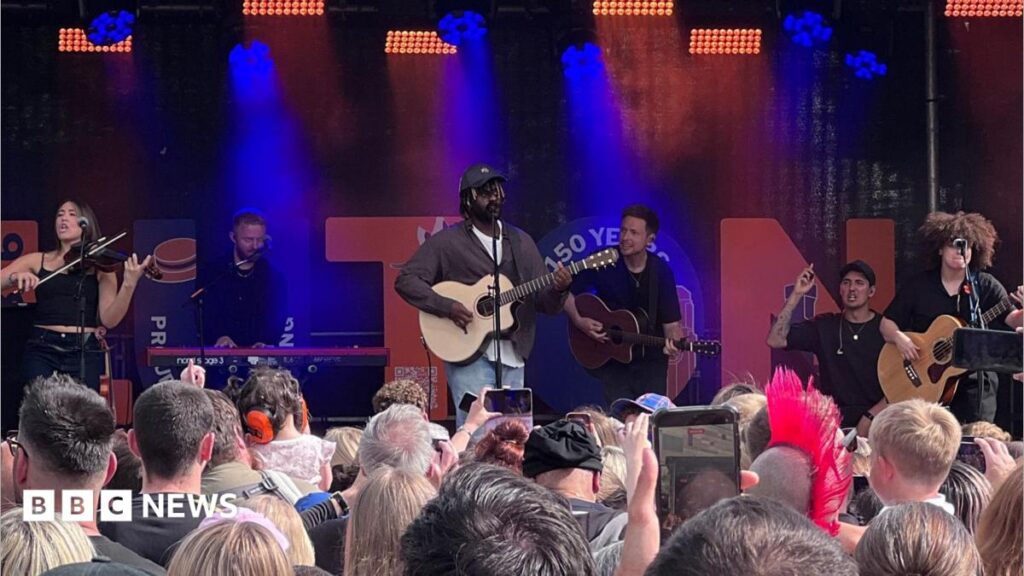 Myles Smith playing a guitar on stage with various other people playing instruments. He is wearing a brown shirt, white T-shirt, jeans and a black baseball cap. A crowd of people are gathered in front of him.