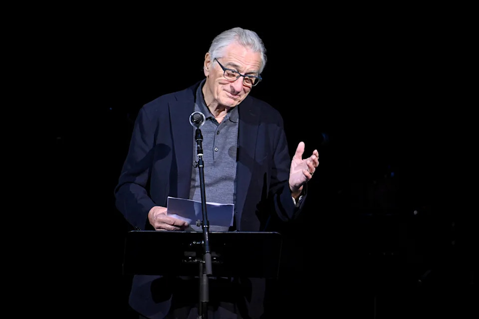 An older man in glasses and a suit jacket speaks into a microphone, holding papers on stage, appearing to give a speech or presentation