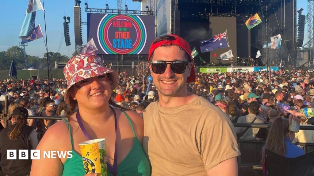A woman and a man stand illuminated by sun in front of a festival stage, with a large crowd watching the stage.