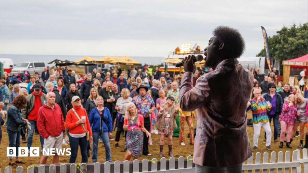 Music festival in Dorset scrapped as organiser vows ‘never again’ A man on stage with black hair and a pink satin jacket sings into a microphone as he looks out into a crowd of people. Behind the audience is a row of stalls and fairground rides and beyond that is the sea.