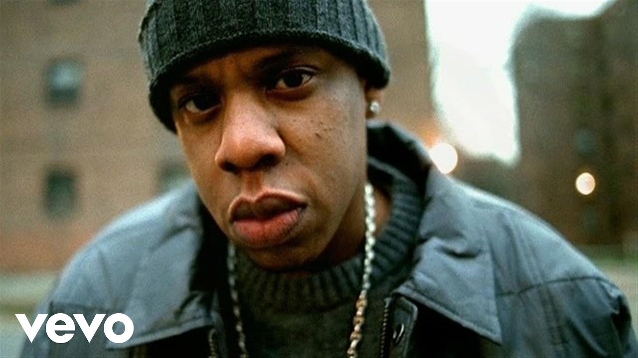 A man wearing a dark knit beanie, gray sweater, leather jacket, and chain necklace gazes seriously at the camera. Out-of-focus brick buildings appear in the background. The "vevo" logo is visible in the bottom left corner.