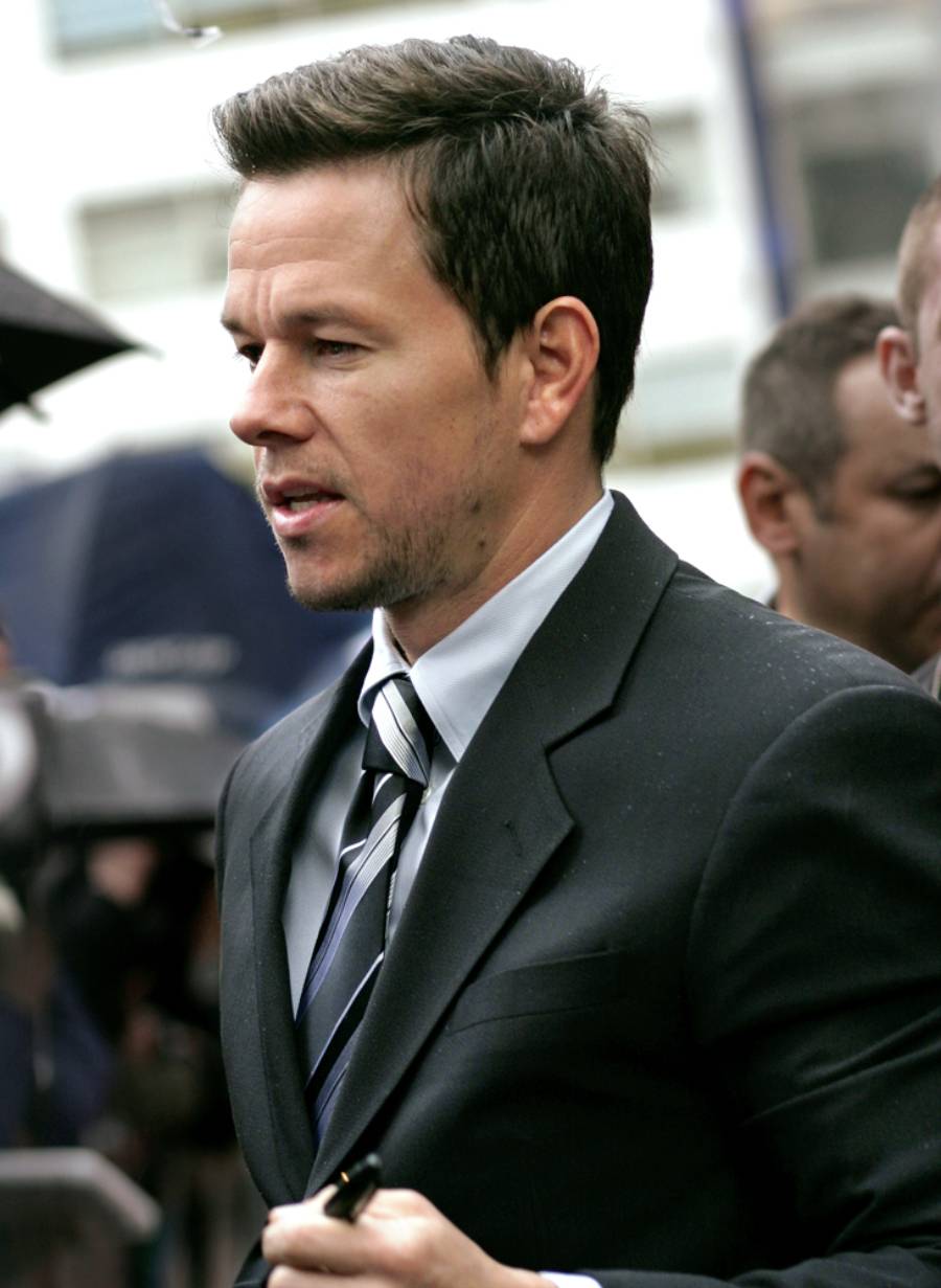 A man in a dark suit and striped tie stands outdoors, holding a pen and appearing to sign something. He has short brown hair and light facial hair, with people and buildings blurred in the background.