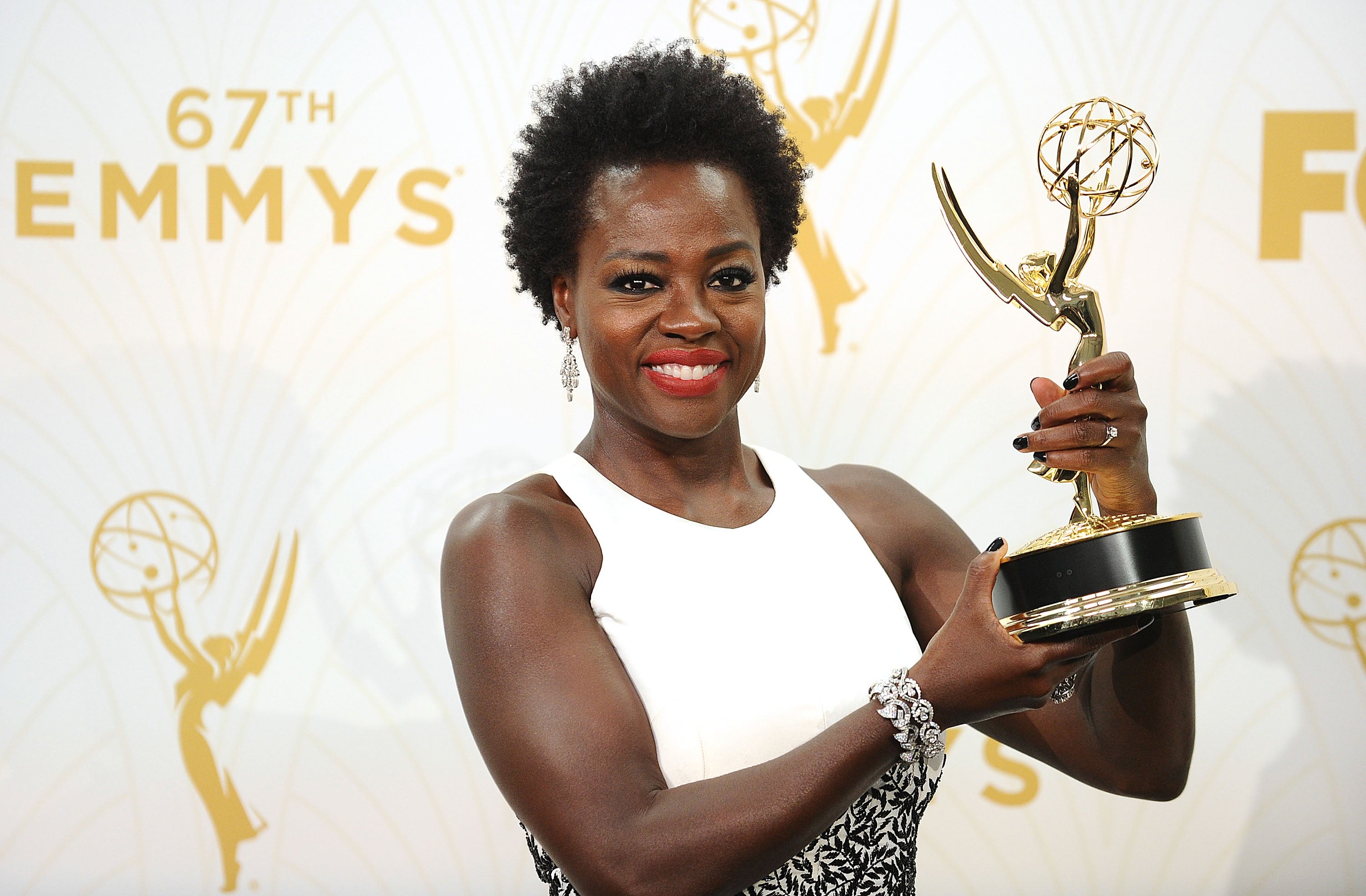 Viola Davis holding an Emmy award, wearing a sleeveless white dress, smiling at an event backdrop with Emmy logos