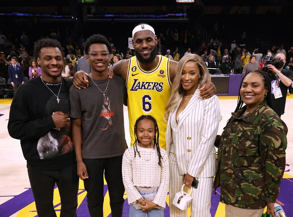 Group of people posing on a basketball court; one wears a Lakers jersey, another in a striped suit, others in casual attire