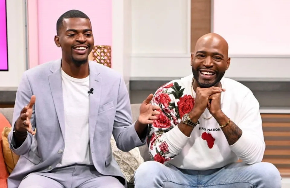 Two men sitting on a couch, smiling and laughing during a talk show. One wears a suit, the other a casual sweatshirt with a floral design