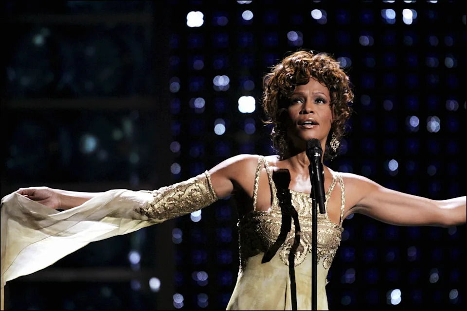 A performer in an elegant, embellished gown sings passionately on stage with outstretched arms, in front of sparkling backdrop