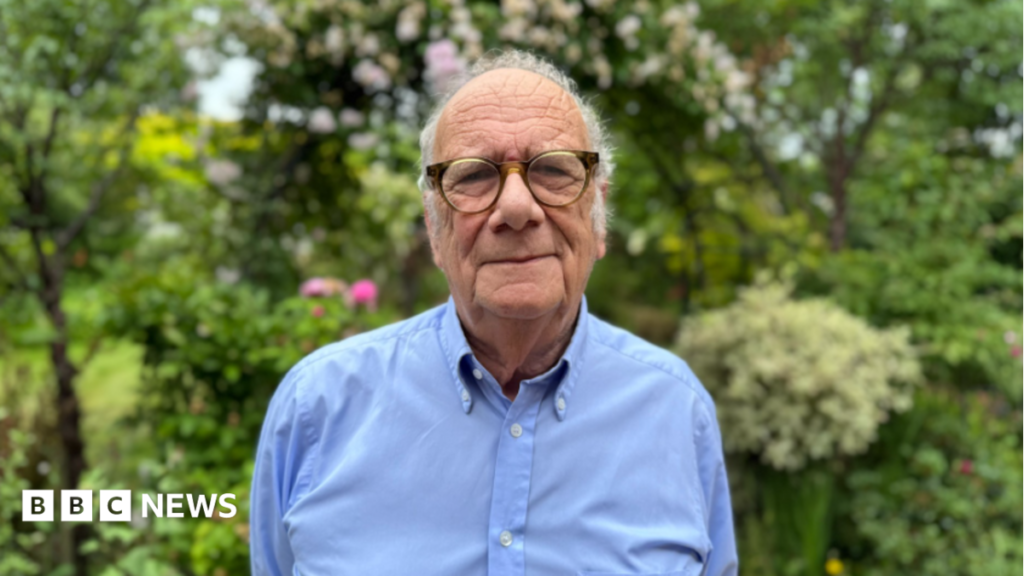 A man with thin grey hair and glasses in a long-sleeved blue shirt in front of green bushes