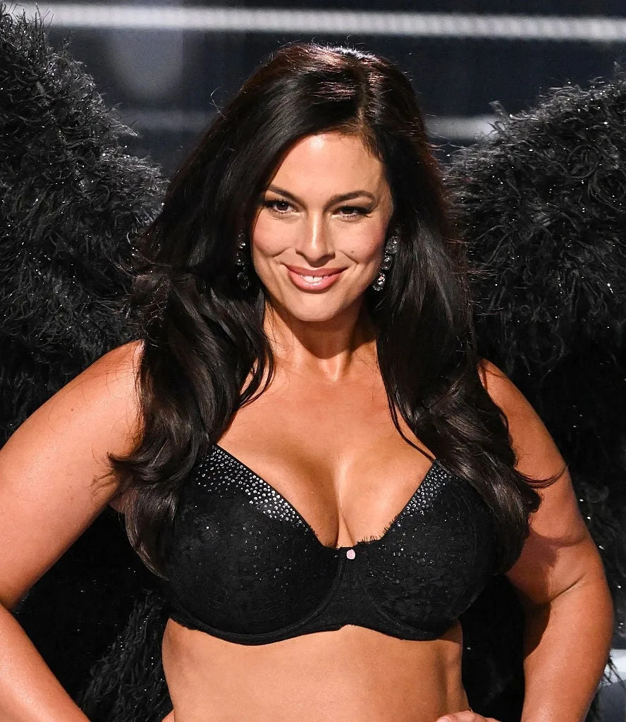 A model wearing black lingerie and feathered wings walks the runway, smiling confidently