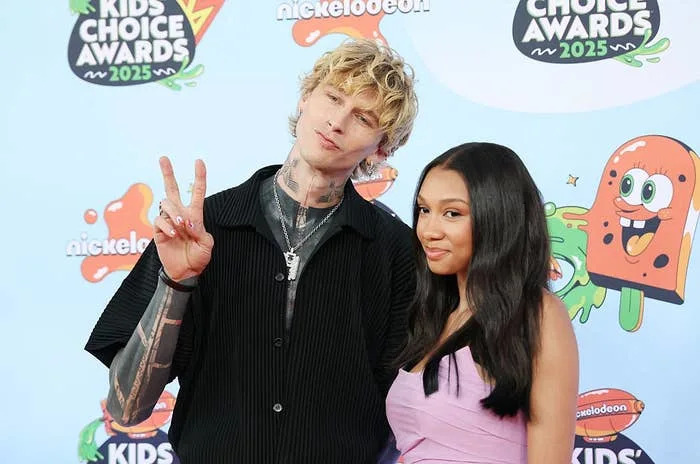Two people on the Kids' Choice Awards 2025 red carpet; one in a casual black outfit giving a peace sign, the other in a stylish purple dress