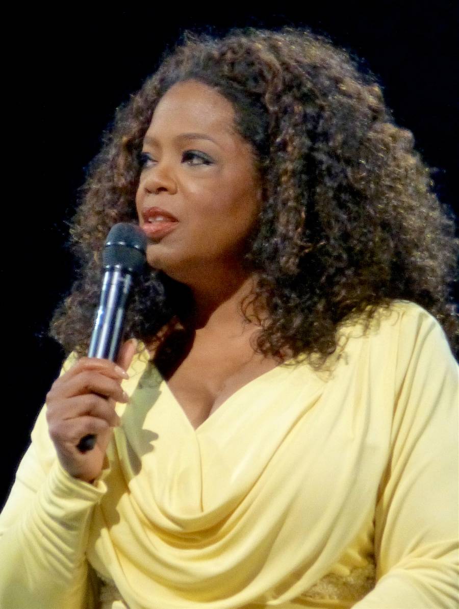 A woman wearing a pale yellow dress holds a microphone and speaks. She has shoulder-length, curly brown hair and is looking to the side against a dark background.