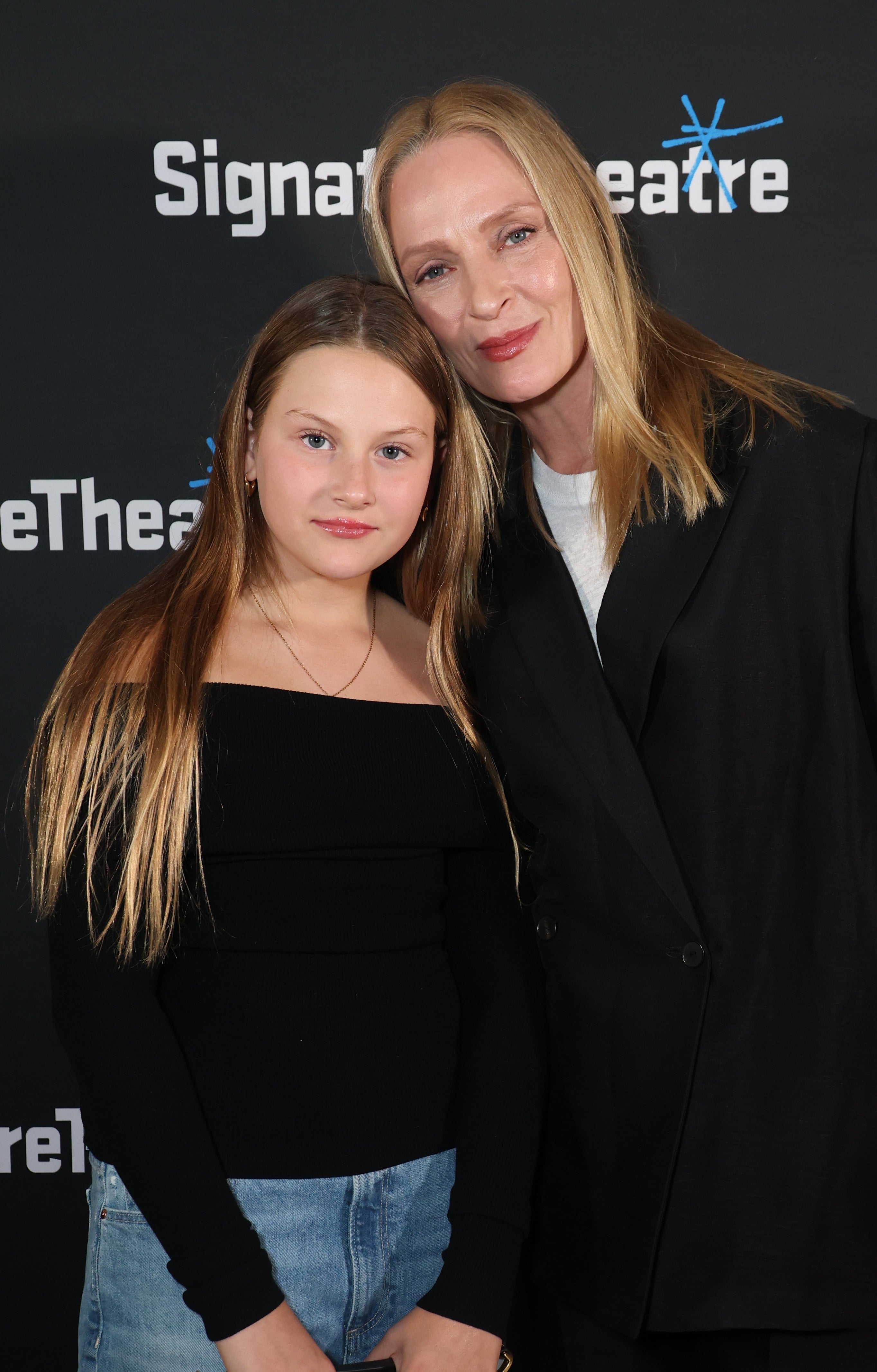 Two individuals pose at a Signature Theatre event; one in a black off-shoulder top and jeans, the other in a black suit