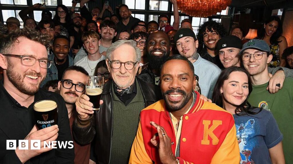 Steven Spielberg makes surprise appearance at London pub quiz Film director Steven Spielberg (C) in a black leather jacket, green jumper and bandana around his neck, poses with fans inside a London pub while holding an half-full glass of Guinness. To his left is actor Colman Domingo in a red and yellow letterman jacket.