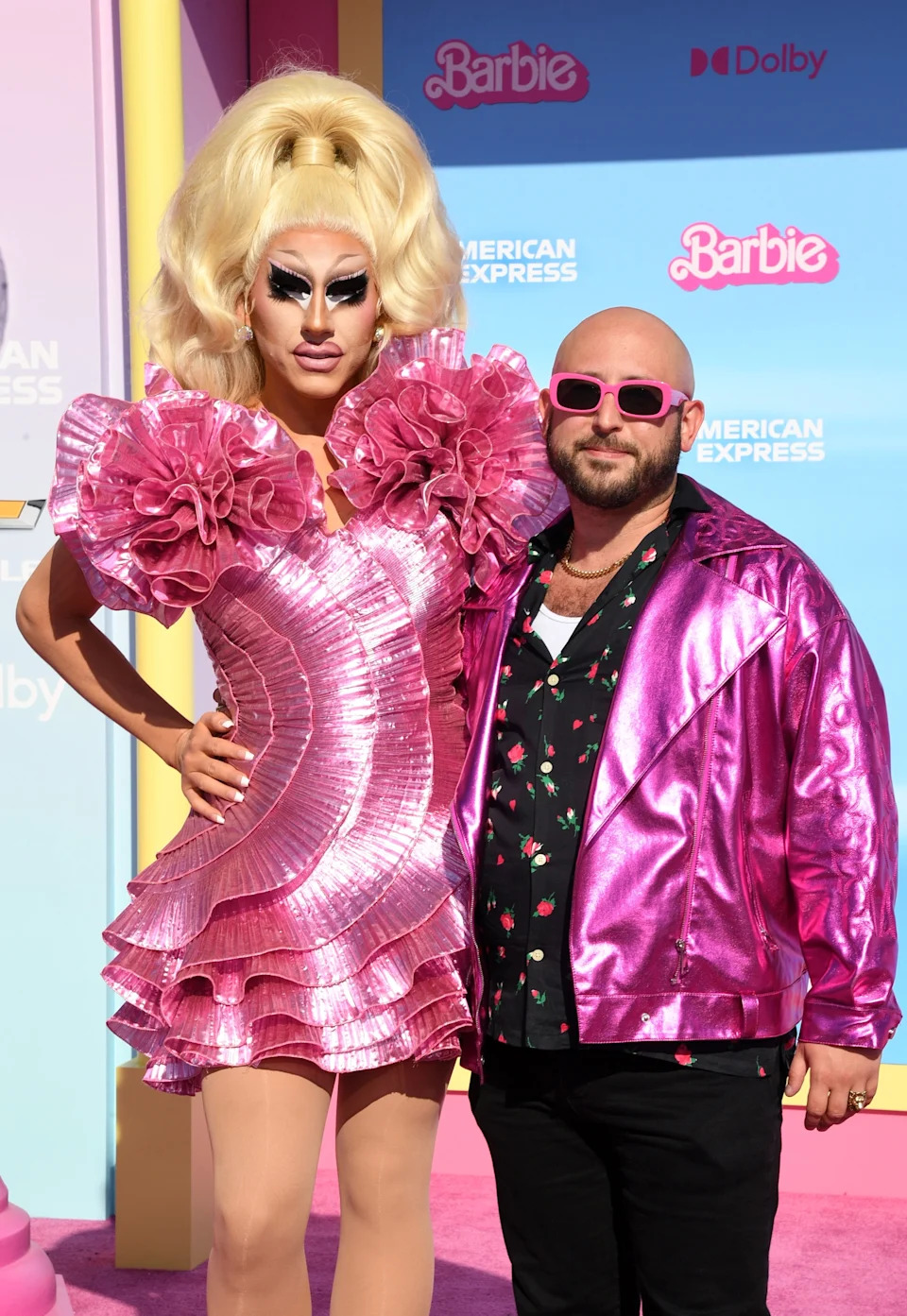 A person in a glamorous ruffled dress stands with someone in a shiny jacket, both posing confidently at an event