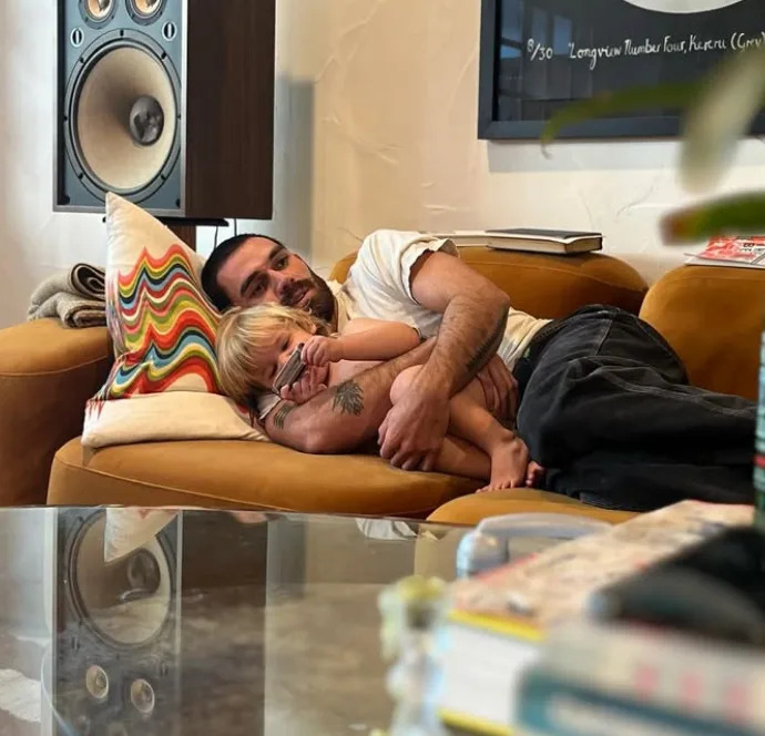 A man and a child cuddle on a sofa in a cozy living room, beside a colorful pillow and large speaker, creating a warm, relaxed scene