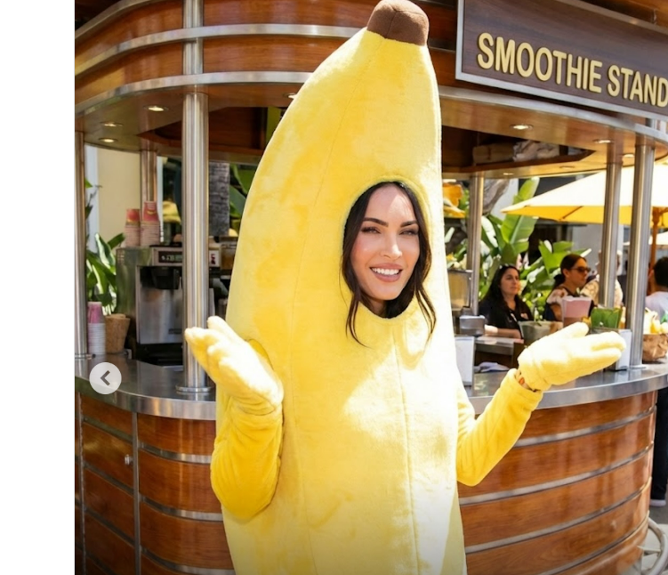 A woman wearing a bright yellow banana costume smiles and gestures with her hands in front of a smoothie stand at an outdoor location. Other people sit at tables in the background.