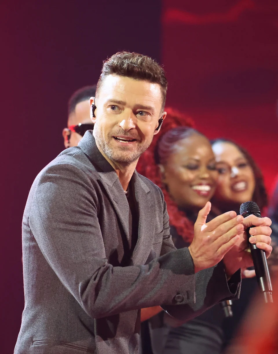 Justin Timberlake in a suit claps on stage, smiling, with background performers