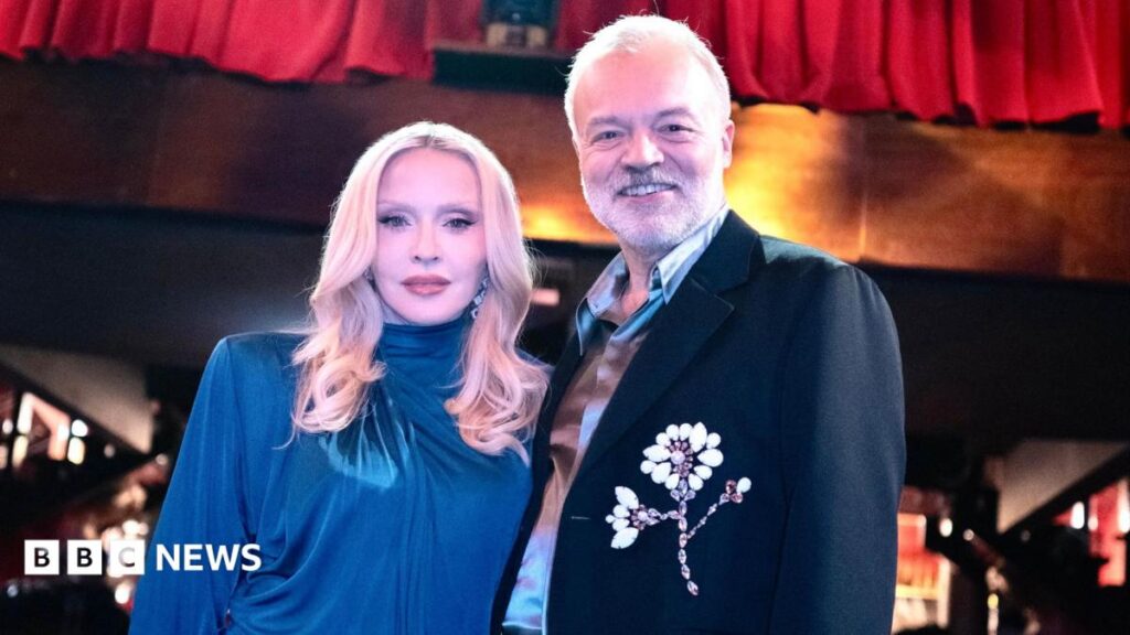 Madonna and Graham Norton are pictured together. She is wearing a blue dress, he is in a dark jacket with a flower motif. They are photographed against a nightclub background.