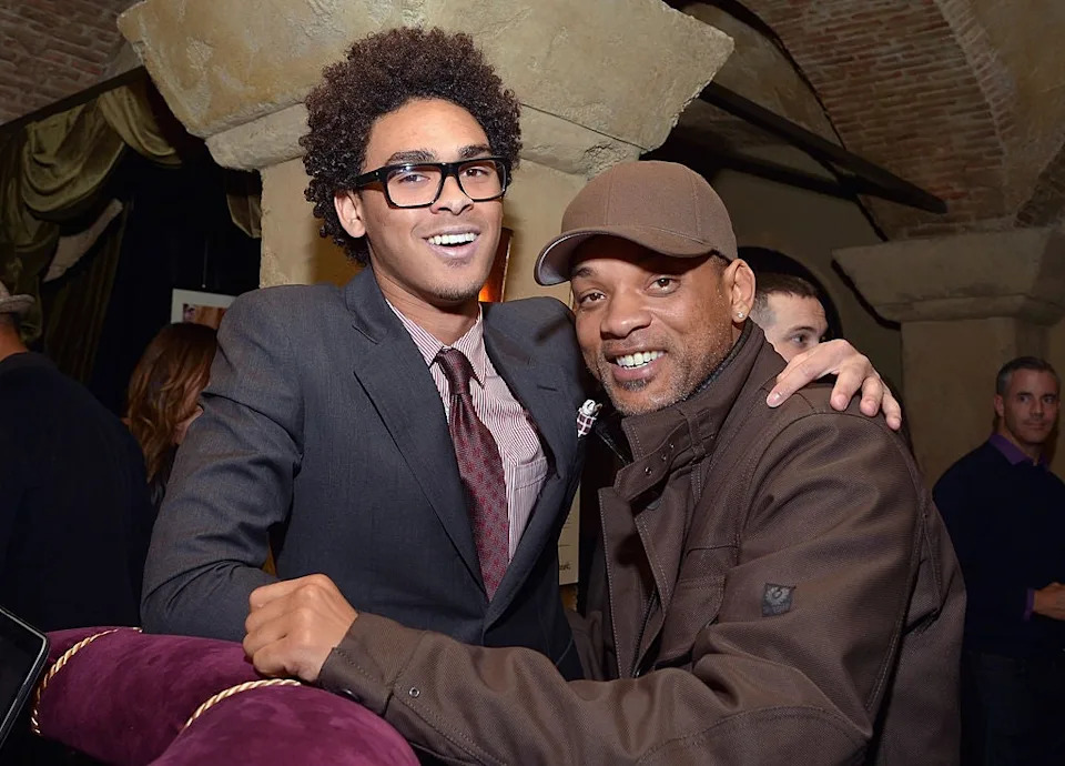 Two people smiling at a social event, one wearing a suit and tie, the other in casual attire with a hat