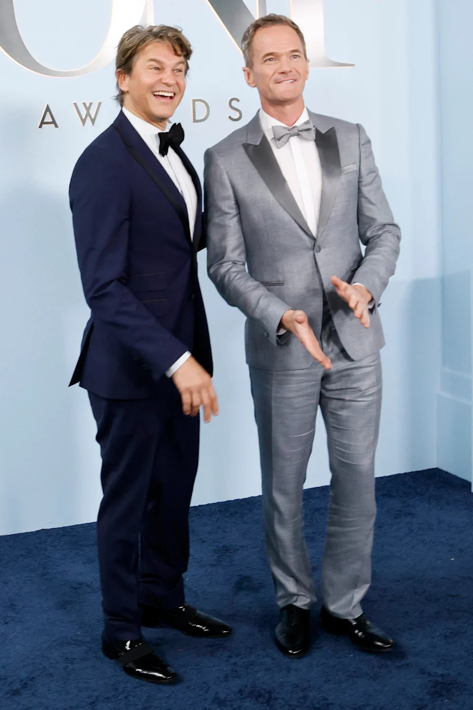 Two individuals in suits stand smiling on a red carpet at an award event; one wears a dark suit, the other a shiny, tailored suit